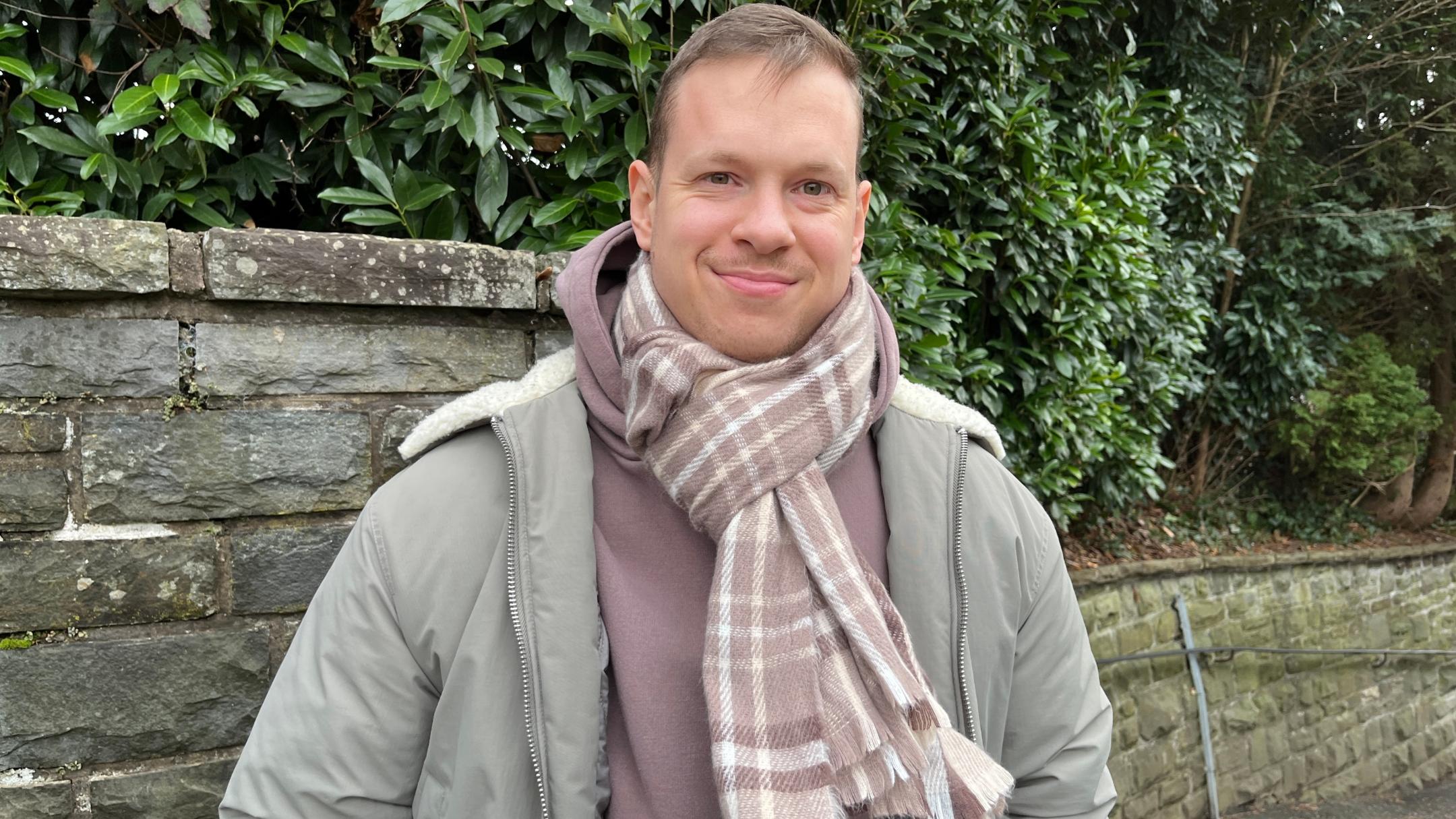 Ein junger Mann mit kurzem, hellbraunem Haar und freundlichem Lächeln steht vor einer Natursteinmauer mit dichten grünen Büschen im Hintergrund. Er trägt eine olivgrüne Winterjacke mit heller Kapuze, einen braunen Kapuzenpullover und einen beigefarbenen karierten Schal, der um seinen Hals gewickelt ist. Seine Hände sind in den Jackentaschen, und er blickt direkt in die Kamera. (Alt-Text erstellt mit Unterstützung von ChatGPT)