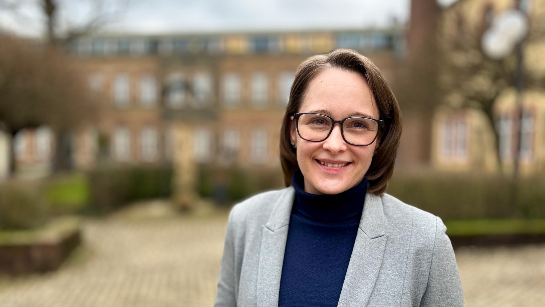 Eine Frau mit schulterlangem braunen Haar und Brille steht lächelnd im Freien vor einem historischen Backsteingebäude. Sie trägt einen grauen Blazer über einem dunkelblauen Rollkragenpullover. Der Hintergrund ist leicht unscharf, mit einem gepflasterten Weg, Hecken und Bäumen.