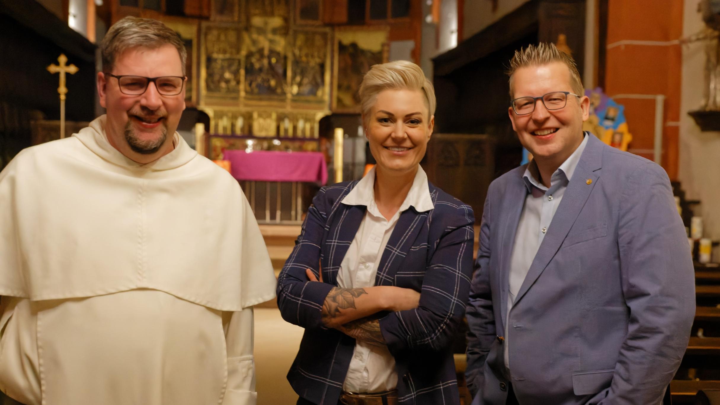 Das Foto zeigt drei Personen in einer Kirche, die freundlich in die Kamera lächeln. Von links nach rechts sind Pater Albert Seul in einer weißen Ordenskleidung, Dr. Carola Holzner aka Doc Caro in einem dunkelblauen karierten Blazer mit verschränkten Armen und Tobias Marenberg in einem hellblauen Anzug zu sehen. Der Hintergrund zeigt einen vergoldeten Hochaltar mit einer violetten Altardecke. (Alt-Text erstellt mit Unterstützung von ChatGPT)