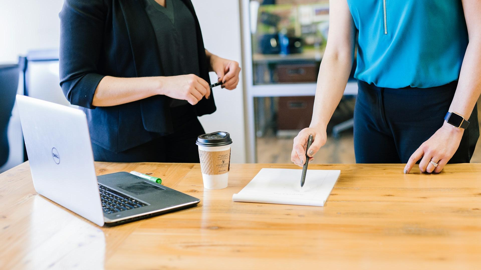 Das Foto zeigt zwei Frauen in einem Büro. Auf dem Tisch liegen ein Laptop, ein Kaffeebecher und ein grüner Marker. Im Hintergrund sind Glaswände und Büromöbel zu sehen.