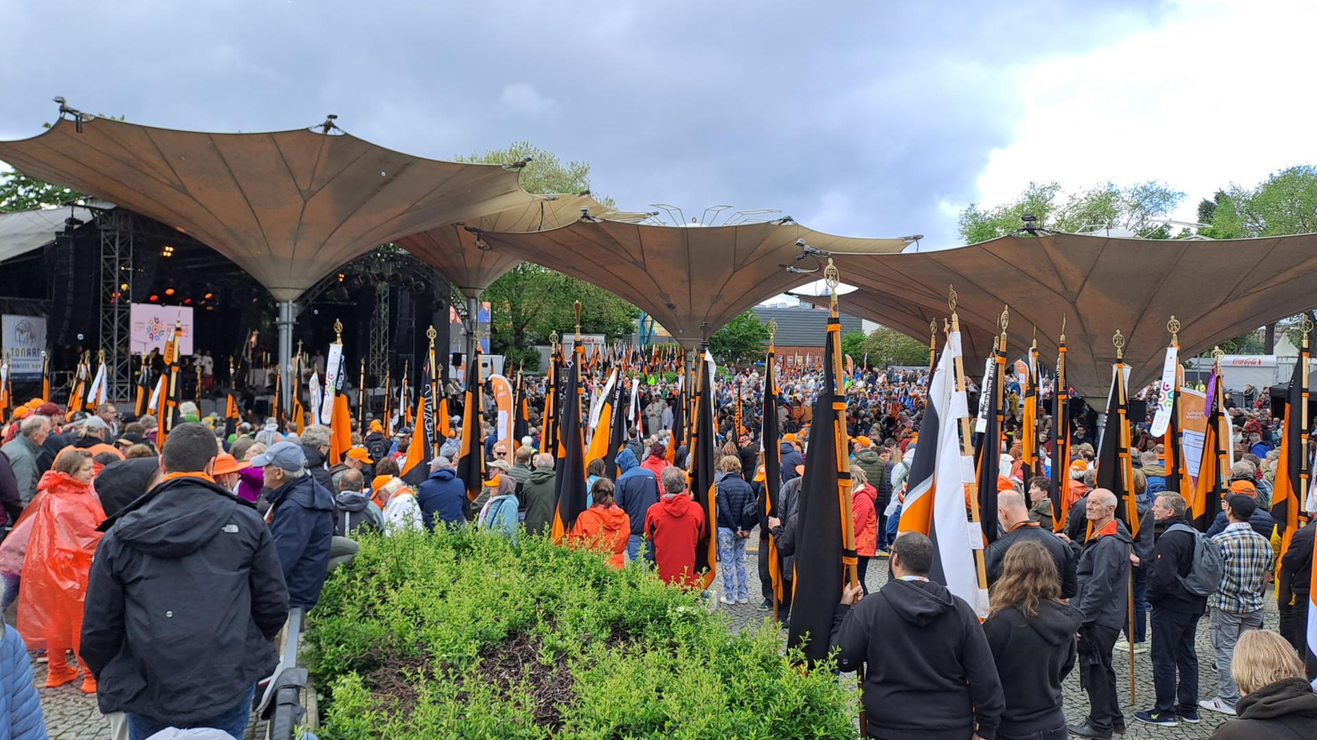 Auf dem Bild ist eine große Menschenmenge bei einer Veranstaltung im Freien zu sehen, viele von ihnen tragen orangefarbene Kleidung oder Accessoires. Zahlreiche orange-schwarze Fahnen mit goldenen Spitzen werden aufrecht gehalten. Im Hintergrund befindet sich eine überdachte Bühne mit Lautsprechern und Bildschirmen unter großen, zeltartigen Dächern. (Alt-Text erstellt mit Unterstützung von ChatGPT)