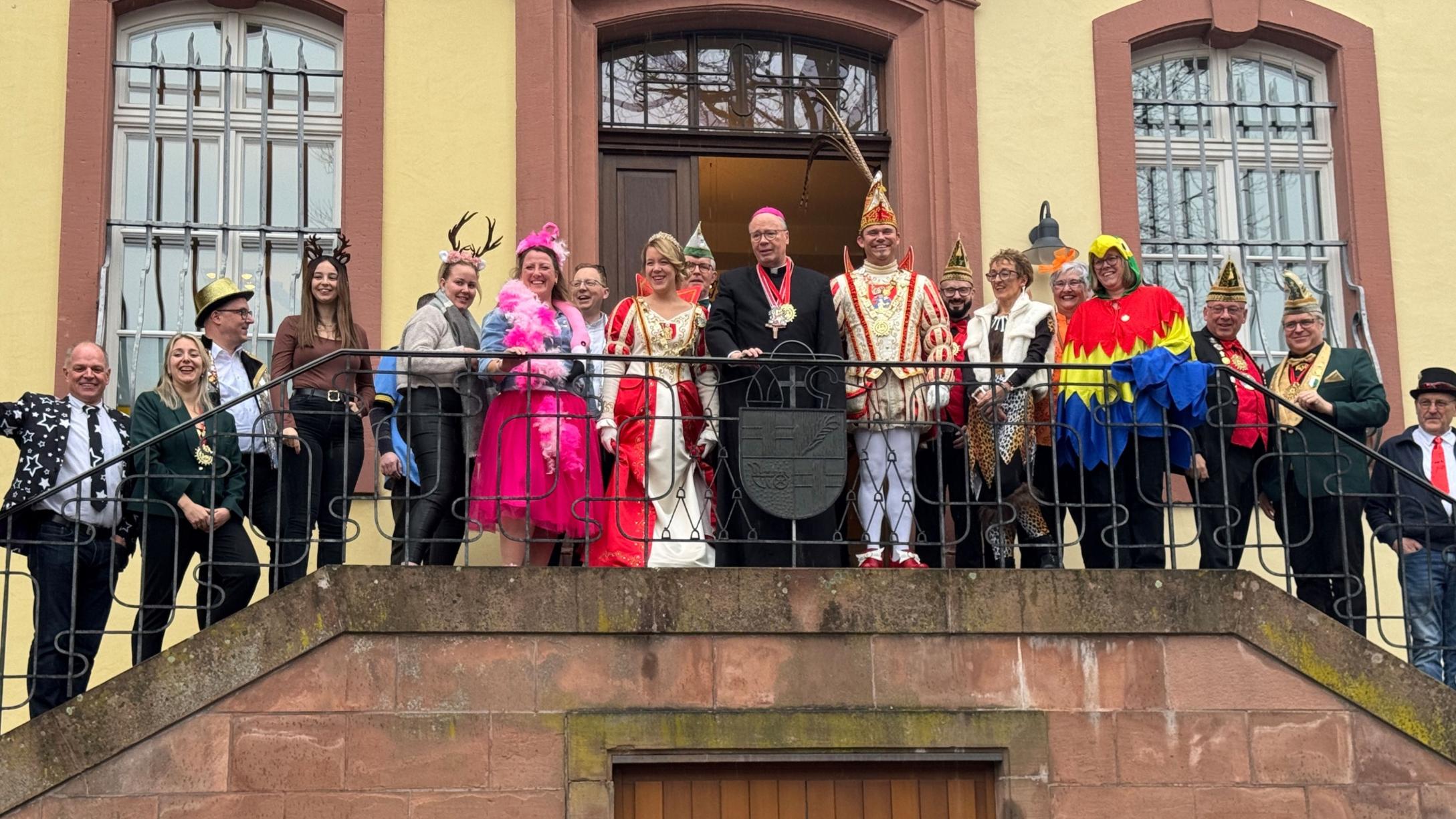Eine große Gruppe steht auf einem Balkongang vor einem historischen Gebäude mit hellen Wänden und hohen Fenstern. In der Mitte befindet sich Bischof Ackermann in dunkler liturgischer Kleidung. Neben ihm stehen das Prinzenpaar in aufwendigen rot‑weißen und rot‑goldenen Karnevalskostümen sowie Angehörige ihres Gefolges mit traditionellen Narrenkappen. Rundherum stehen Mitarbeitende des Bischofshofes und des Generalvikariates.