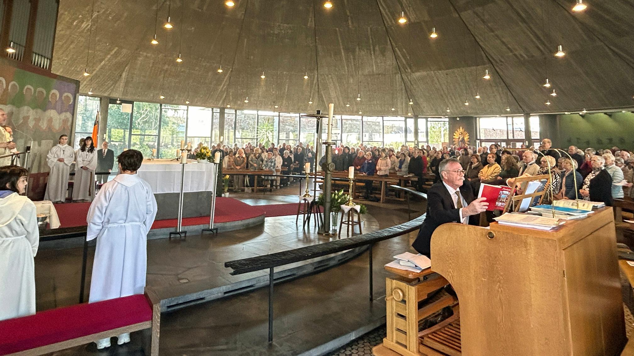 Innenaufnahme einer großen, modern gestalteten Kirche während eines festlichen Gottesdienstes. Die Gemeinde füllt den gesamten Raum, viele Personen sitzen oder stehen dicht beieinander in den Bankreihen, die halbkreisförmig um den Altar angeordnet sind. Der Altar ist zentral positioniert, weiß gedeckt und von liturgischen Diensten umgeben, darunter mehrere Personen in hellen liturgischen Gewändern. Im linken Bereich stehen weitere Personen im Altarraum und beteiligen sich am Gottesdienst. Rechts im Vordergrund sitzt eine Person an einer großen Orgel und spielt, vor sich Noten und Bücher auf dem Instrument. Der Kirchenraum ist lichtdurchflutet durch hohe Fenster im Hintergrund, und zahlreiche kleine Lampen hängen von der dunklen, zeltartigen Decke herab. Insgesamt vermittelt das Bild den Eindruck eines feierlichen und gut besuchten Festhochamts.