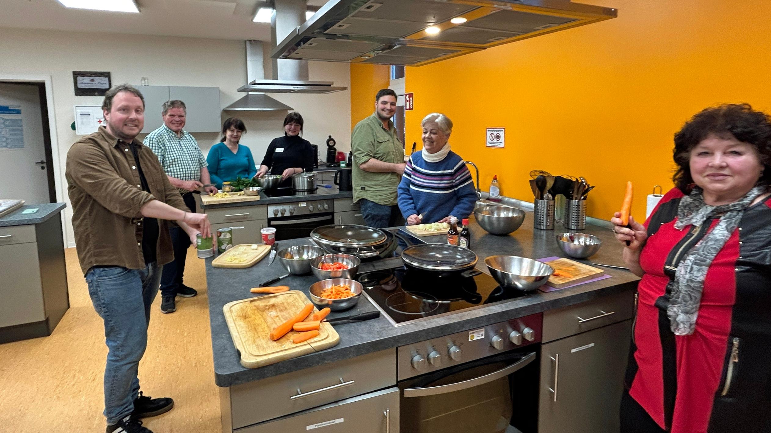 Eine Gruppe von sieben Menschen steht in einer Lehrküche und bereitet gemeinsam ein Essen zu. Mehrere Personen schneiden Gemüse, darunter Karotten, und lächeln in Richtung Kamera. Auf der zentralen Arbeitsfläche liegen Schneidebretter, Messer, Schüsseln und Pfannen mit bereits geschnittenem Gemüse. Die Küche ist modern ausgestattet, mit Edelstahlflächen, Kochfeldern und einer leuchtend orangefarbenen Wand im Hintergrund. Die Stimmung wirkt freundlich und gemeinschaftlich.