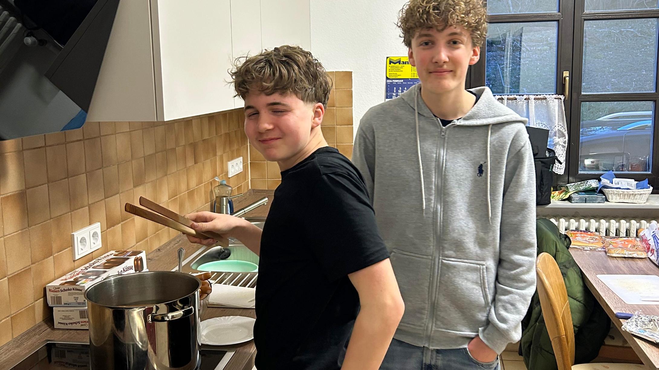 Zwei Jugendliche stehen in einer Küche. Eine Person steht am Herd und rührt mit Küchenzangen in einem großen Edelstahlkochtopf auf der Arbeitsplatte. Daneben liegen ein Topfdeckel und Zutaten wie Hotdog-Brötchen. Die zweite Person steht leicht seitlich dahinter, mit den Händen in den Hosentaschen. Im Hintergrund sind Küchenschränke, ein Fenster, ein Esstisch mit Stühlen und weitere Lebensmittel zu sehen.