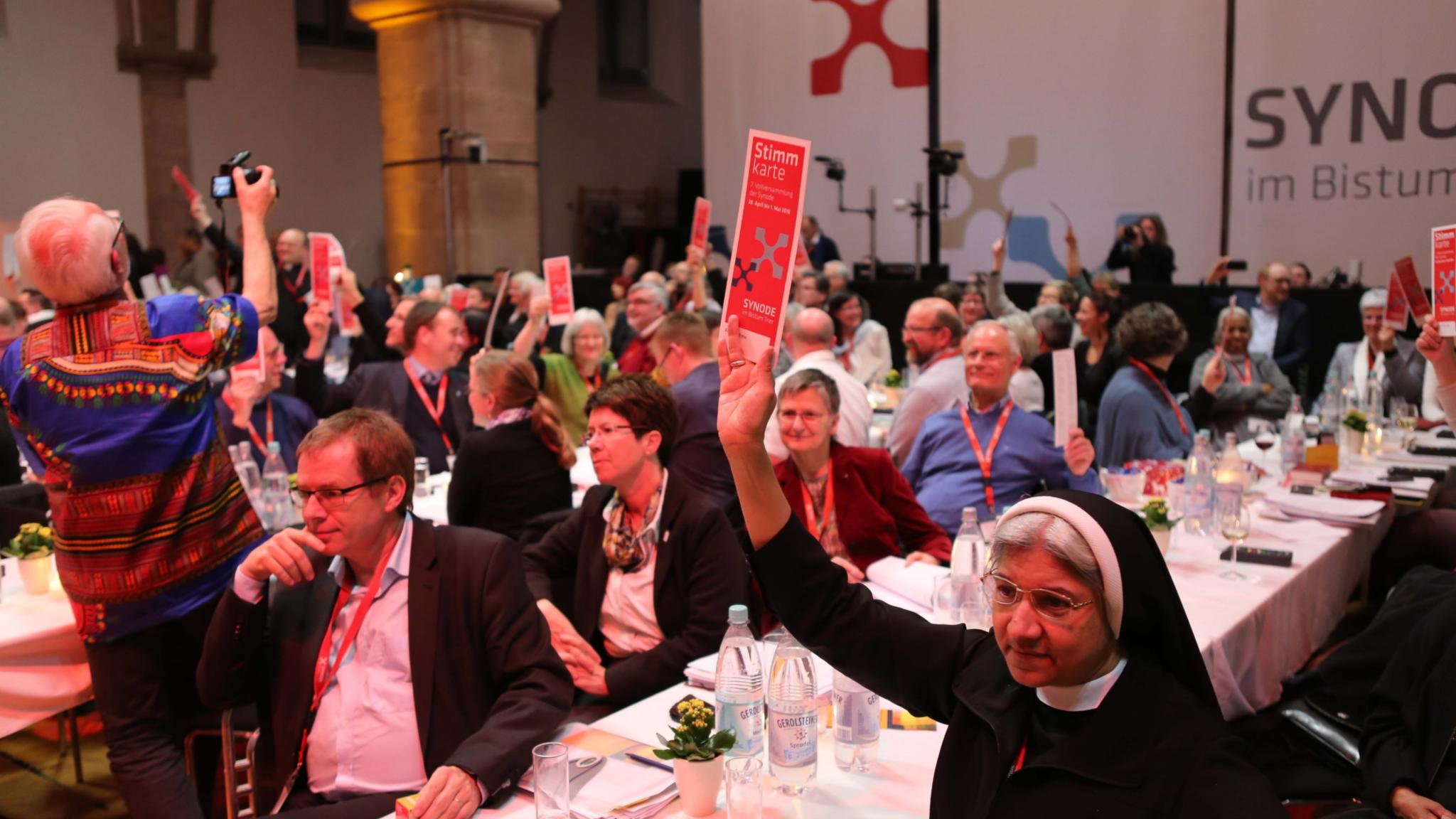 Auf dem Bild sind zahlreiche Menschen in einem großen Saal bei einer Veranstaltung zu sehen, viele von ihnen halten rote Karten mit der Aufschrift „Stimmkarte“ in die Höhe. Im Vordergrund sitzt eine Ordensschwester, die ebenfalls eine Stimmkarte hochhält, während andere Teilnehmer aufmerksam blicken oder diskutieren. Im Hintergrund ist ein großes Transparent mit der Aufschrift „Synode im Bistum“ sichtbar. (Alt-Text erstellt mit KI-Unterstützung)