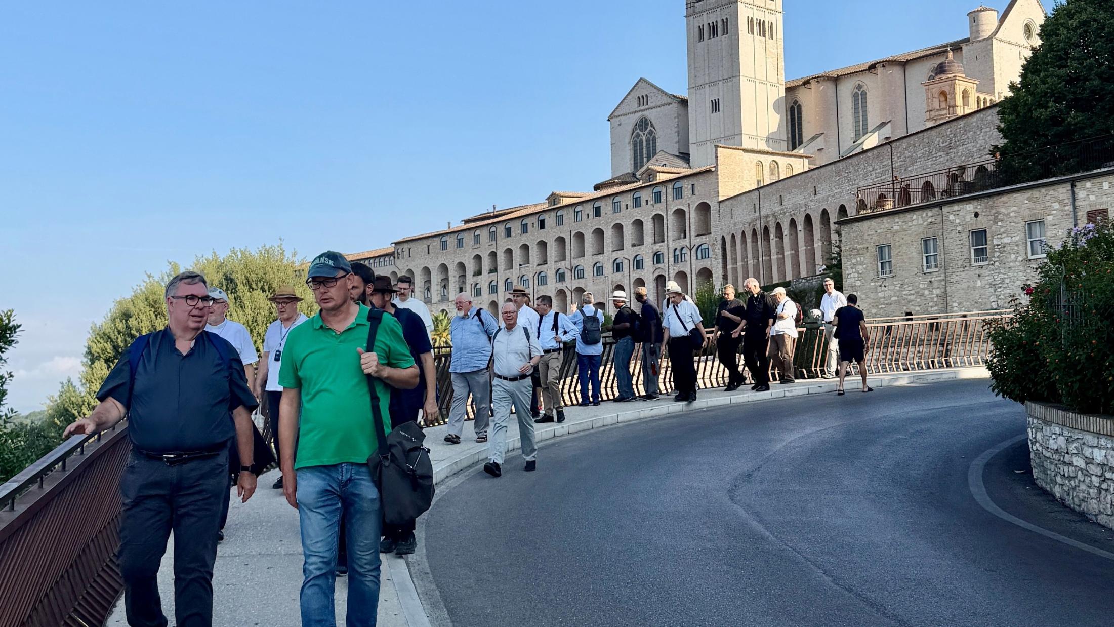 Priester spazieren hintereinander auf einem Gehweg, dahinter die Kirche von Assisi