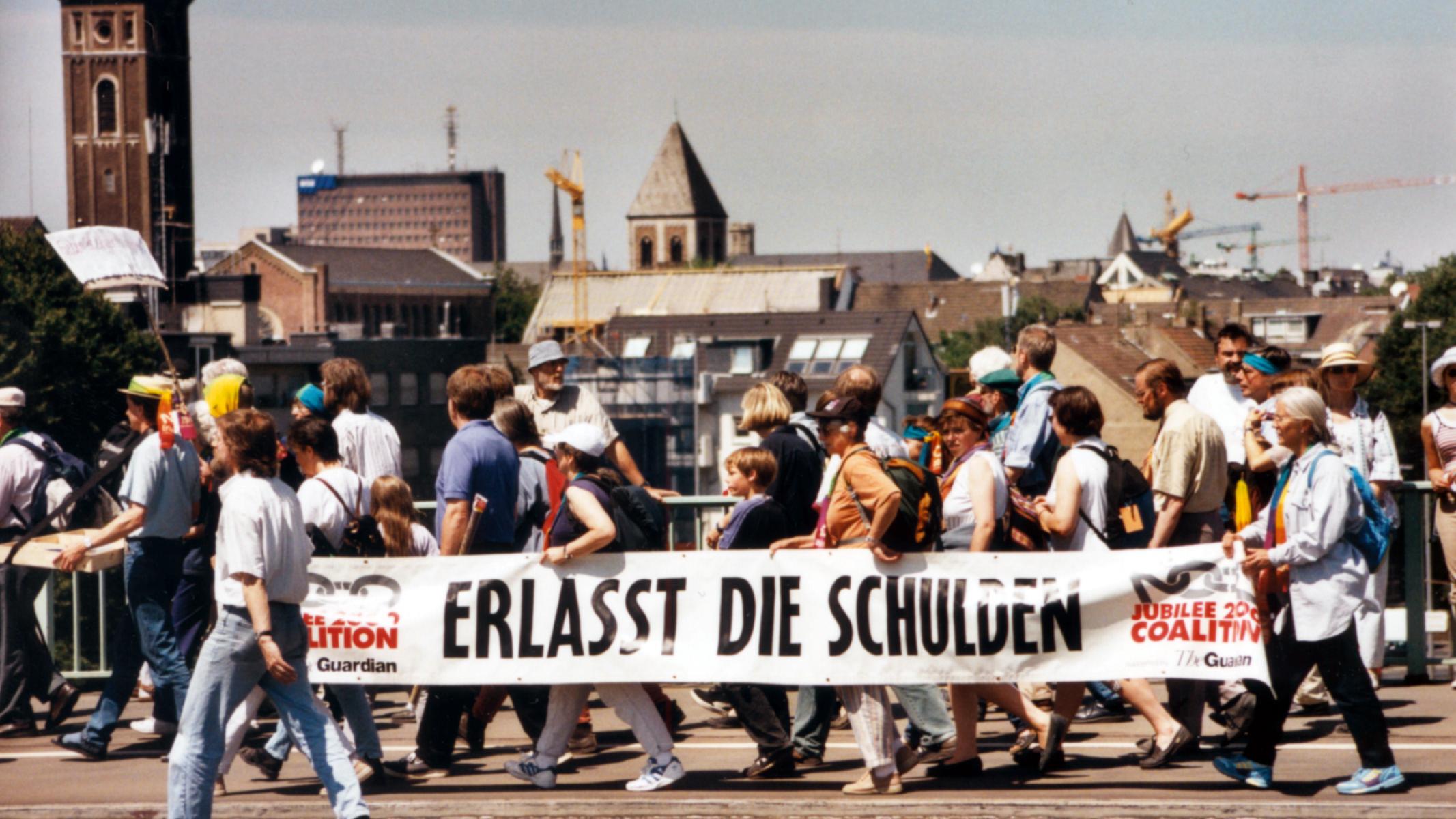 Menschen bei einer Protestaktion tragen ein großes Banner mit der Aufschrift 'Erlasst die Schulden'