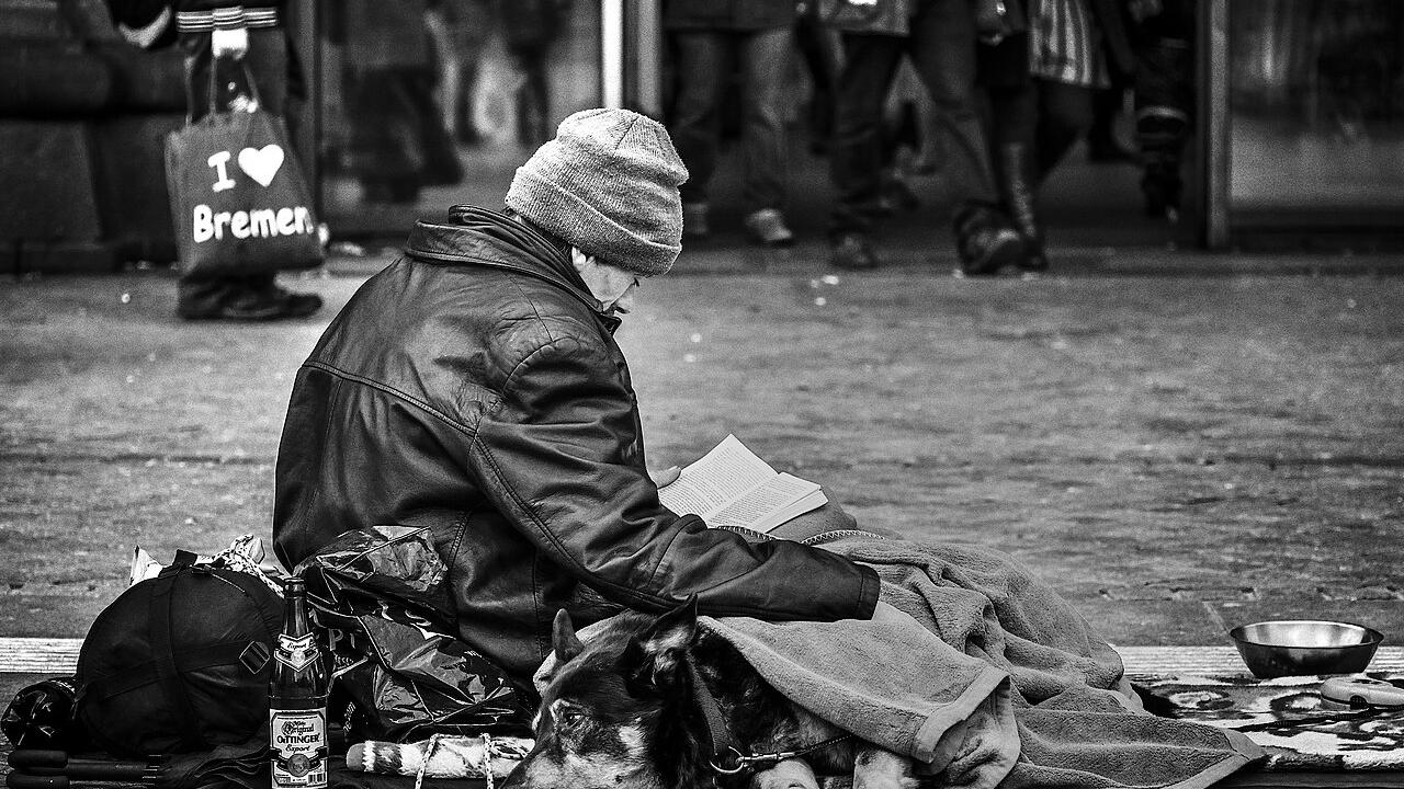 Obdachlose Person sitzt auf Straße, Hund liegt zugedeckt neben ihr.