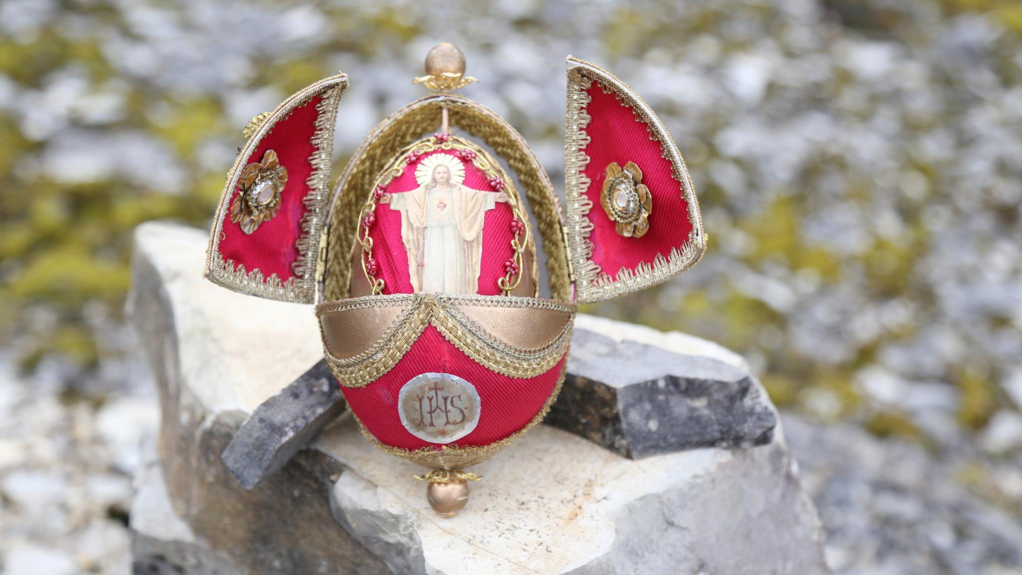Ein verziertes, rot‑goldenes Osterei steht geöffnet auf einem großen Stein in einer felsigen Umgebung. Die beiden seitlichen Klappen des Eis sind nach außen aufgeklappt und mit goldenen Stickereien geschmückt. Im Inneren des Eis befindet sich eine kleine, hellfarbene Figur, die mit ausgebreiteten Armen dargestellt ist. Auf der Vorderseite des Eis ist ein rundes Emblem mit den Buchstaben ‚IHS‘ angebracht.