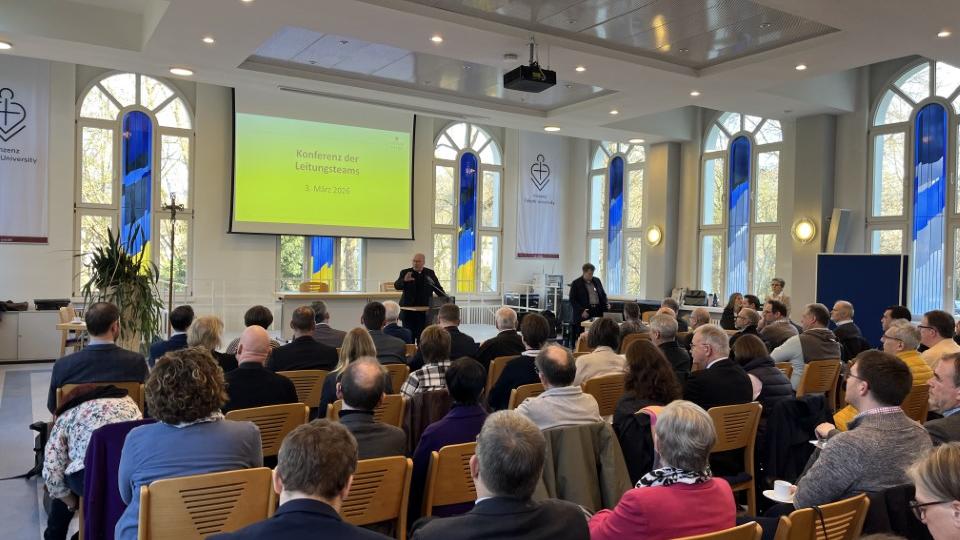 Man sieht viele Menschen, die in einem Saal sitzen und eine Präsentation anschauen