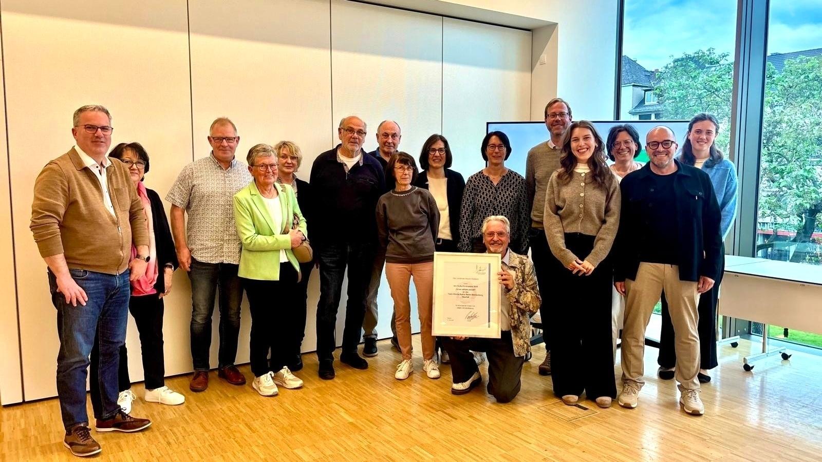 Ein Gruppenfoto mit unterschiedlichen Menschen, in der Mitte hält ein Mann eine Urkunde hoch.