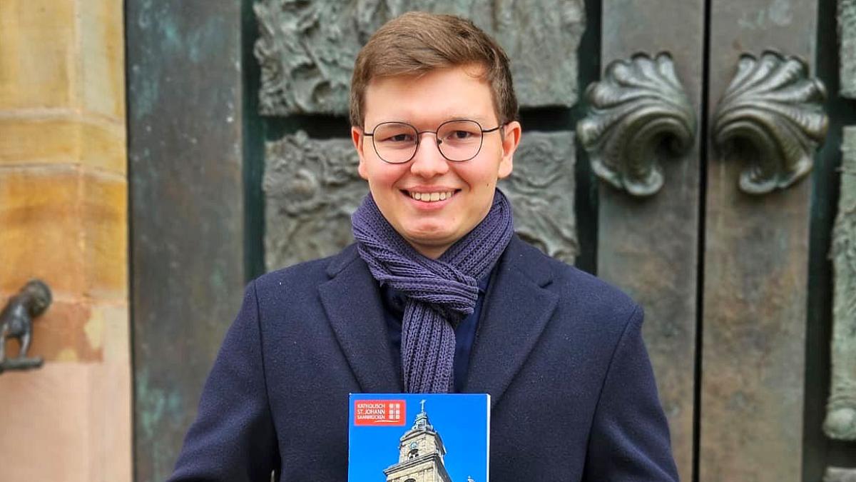Der 19-jährige  David Sawas mit seiner Festschrift über die Saarbrücker Basilika St. Johann vor dem Portal der Kirche, das die künstlerische Handschrift des Bildhauers Ernst  Alt trägt.