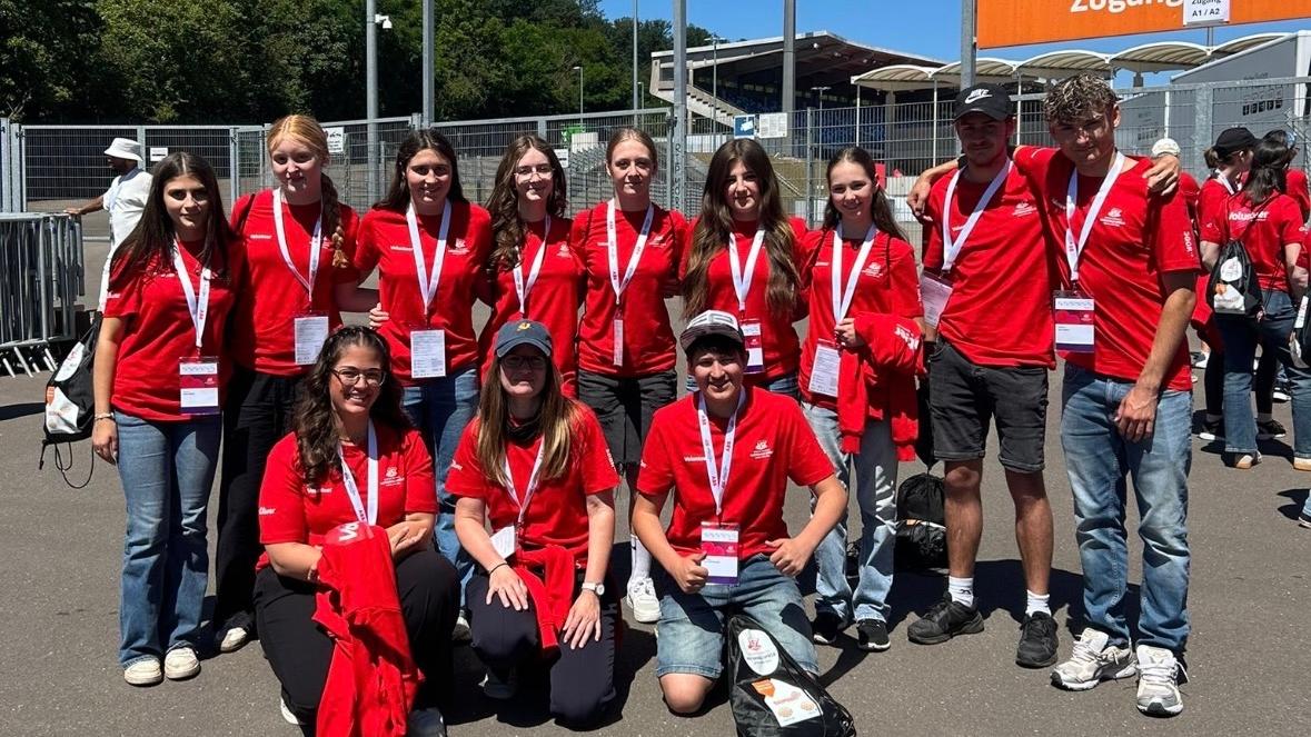 Gruppe von etwa elf Freiwilligen in roten T-Shirts mit Namensschildern und Akkreditierungen vor einem Stadioneingang. Die Personen stehen und knien für ein Gruppenfoto auf einem asphaltierten Platz. Im Hintergrund sind Zäune, Tribünen, Flutlichtmasten und orangefarbene Schilder mit den Aufschriften „Zufahrt Gäste-Fanbusse“ und „Zugang Gästefans“ zu sehen. Sonniger Tag mit klarem blauem Himmel.