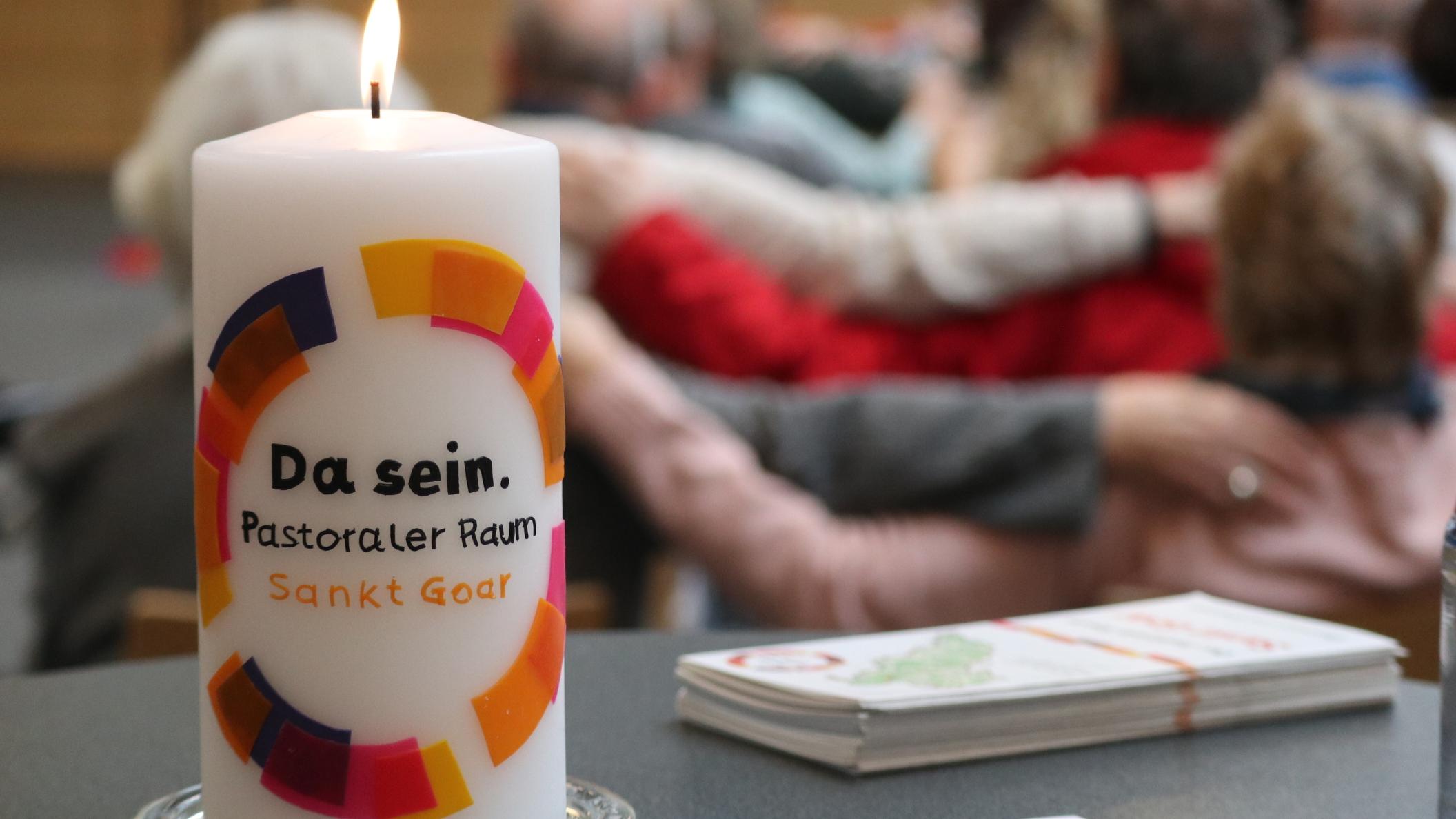 Eine Kerze mit der Aufschrift 'Da sein' steht auf einem Tisch, im Hintergrund sieht man Menschen, die Arme aufeinander legen.