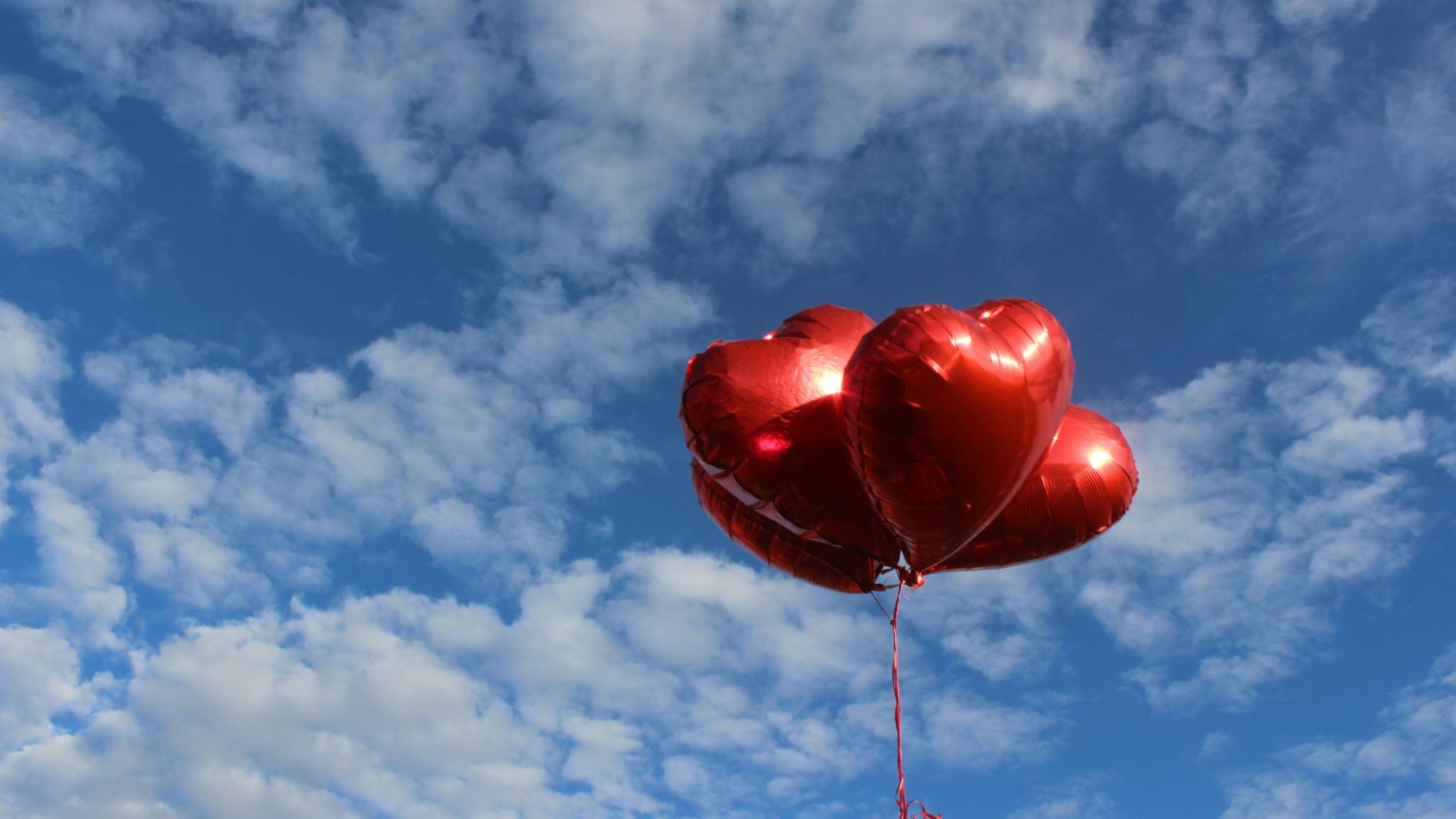 Vor einem blauen Himmel mit verstreuten weißen Wolken schweben drei glänzende, rote, herzförmige Ballons. Sie hängen an dünnen roten Schnüren und heben sich deutlich vom Himmel ab.