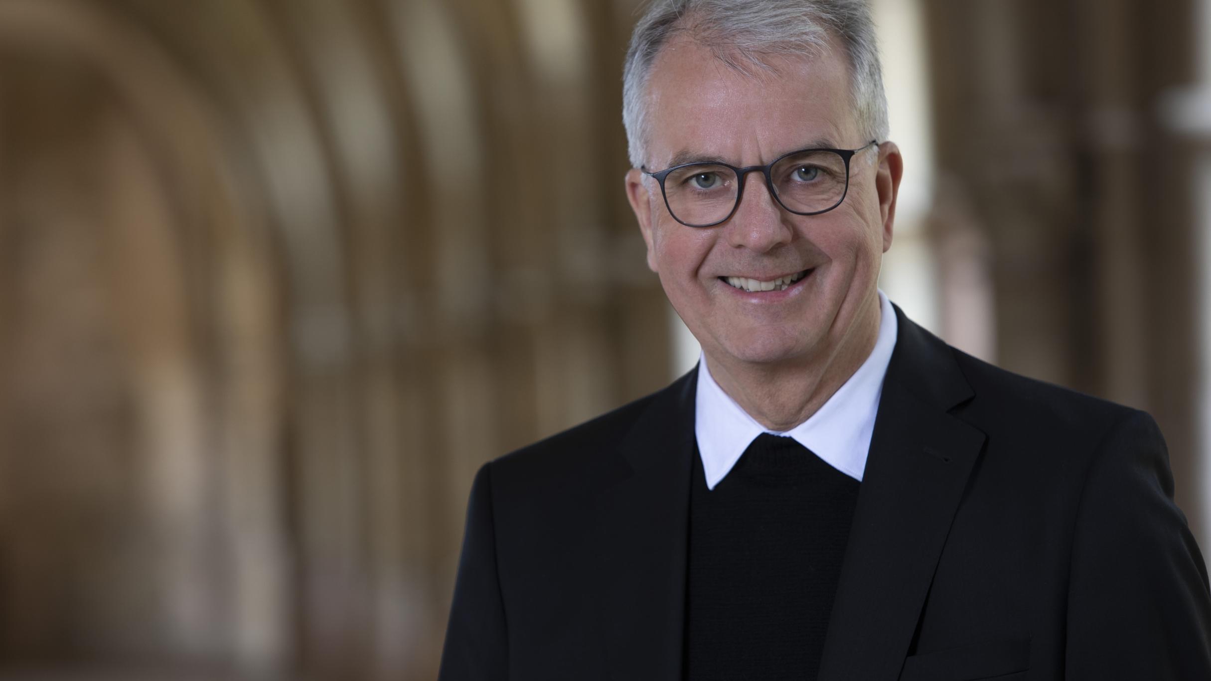 Person im dunklen Anzug in einem Kreuzgang in warmem Licht.