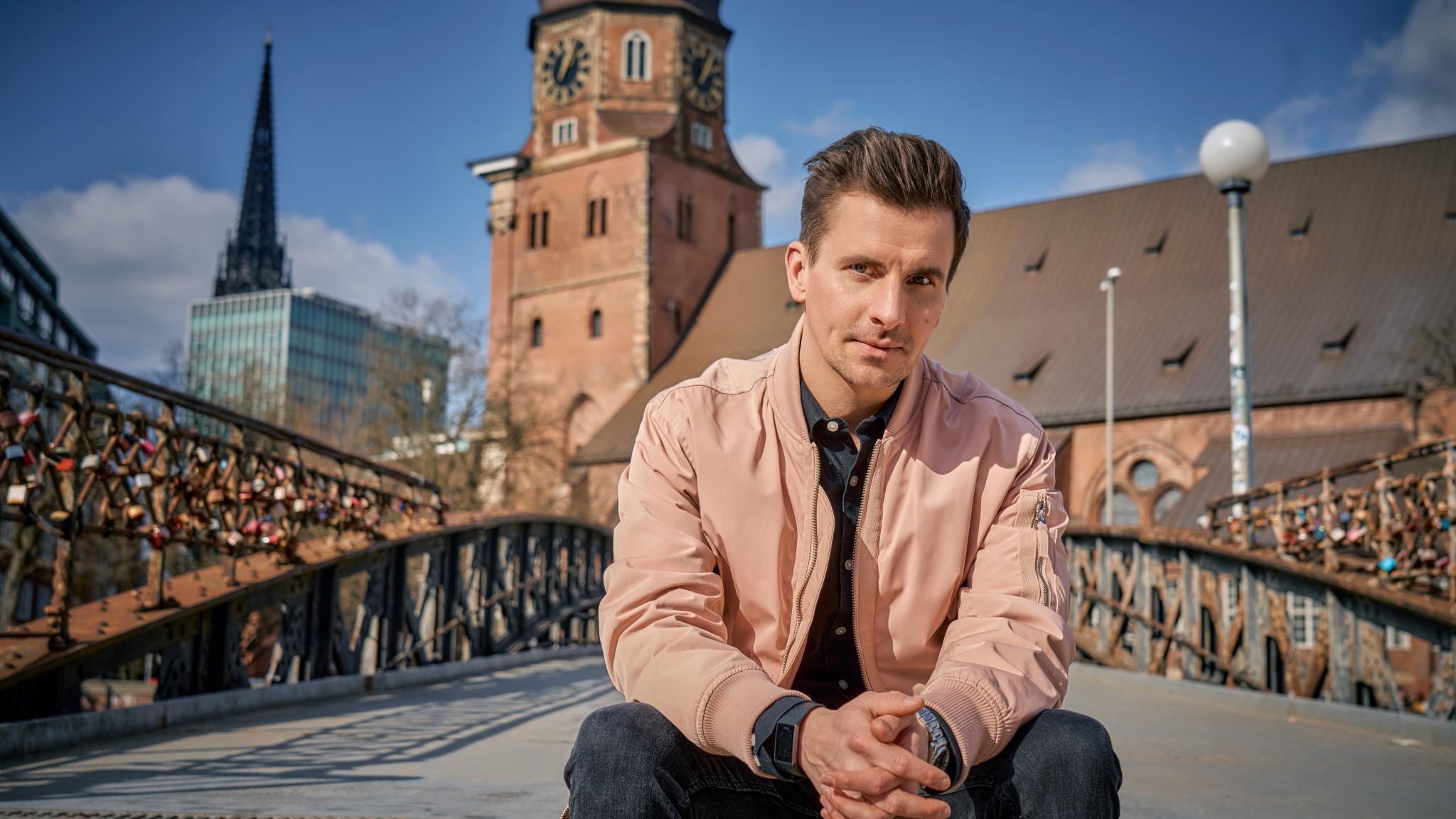 Mann sitzt auf einer Brücke mit Liebesschlössern, Kirche im Hintergrund.
